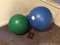 Large blue and medium green exercise balls with a pair of small black dumbbells on carpeted floor, near wall outlet and cabinet.