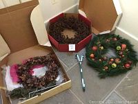 Three door wreaths: one large artificial pine garland wreath with decorative apples, berries, and pine cones; one miniature pine cone wreath inside a cardboard box; one pine cone and acorn wreath wrapped partially in tissue paper in another box; two metal door hangers placed on the floor.