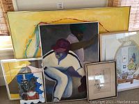 Five framed paintings and drawings grouped together leaning against a wall showing various sizes and themes