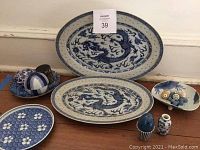 Two large blue and white oval platters with dragon designs, three smaller decorative plates, a blue ceramic sphere, two blue and white jewelry boxes, and a small vase arranged on floor against a wall.
