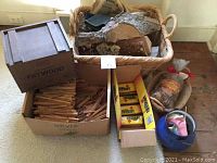 Assorted kindling and logs arranged in wicker basket, box of fire kindling sticks, yellow box of Duraflame fire starter logs, and blue potpourri container on floor