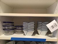 Shelf view showing 6 glass round bowls with blue rim and 6 white ceramic scalloped bowls with blue rim stacked.