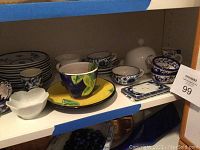 Wide view of assorted blue and white saucers, cups, white butter dish, and colorful cup/saucer set on shelf.