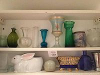 Shelf with assorted glass vases in green, clear, blue, turquoise colors, rabbit planter, small ceramic sphere, and blue and gold decorative basket.