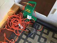 Green electric Weed Eater Ground Sweeper Blower with orange extension cord and black nozzle attachment on floor