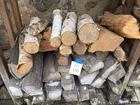 Pile of firewood showing white birch pieces and mixed hardwood logs stacked under a wooden shelter against stone wall.