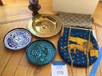 Photo showing all five items together: two ceramic plates, one brass plate, lion needlepoint purse, and tan geometric clutch purse