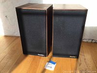 Front view of the two Paradigm speakers side by side on wooden floor with black grills and Paradigm logo on bottom right.