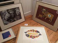 Four framed artworks placed on wooden floor. Right side shows colorful stylized cow painting framed, center foreground shows Fishbread and Corn painting by Jonas dos Santos, left rear shows photo of three statues framed, and front left shows small abstract blue and red art.