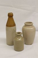 Three vintage stoneware jars and bottles including Virol jar, Cook's Nips crock bottle, and Ginger Beer bottle displayed against white backdrop.