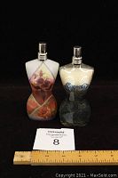 Two vintage Jean-Paul Gaultier perfume bottles shaped like female torsos with decorative dress patterns, placed side by side with measuring ruler and lot tag visible.