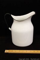 Single large white enamel watering jug with black handle and rim shown against dark background with ruler for scale.