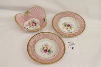 Three pink reticulated porcelain pieces on a white background: one heart-shaped basket bowl with rose decoration and two round ashtrays with floral and gold detailing.