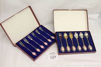 Two open boxes showing 6 silver handled forks in one box and 6 silver-handled spoons in another box with blue velvet lining.