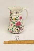 Front and side view of the ceramic pitcher showing the floral and bird pattern, handle, and scalloped rim with green trim against a white background, with a measuring ruler for scale.