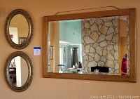Two oval mirrors with ornate gold frames hung vertically on a wall next to a large rectangular wooden framed mirror showing a stone fireplace reflection.