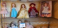 Five Madame Alexander dolls on a metal shelf, including Alice in Wonderland in original box, Wendy, Sleeping Beauty, Molly, and Renoir dolls, showing overall collection.