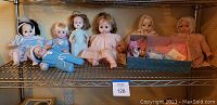 Seven Madame Alexander dolls displayed on a metal shelf, with various doll sizes and clothing, one box with doll accessories.