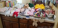 Wide view of miscellaneous doll clothes, hats, and accessories spread on a counter, some hanging below, with a basket containing dolls at far right.