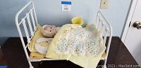 Full view of white iron doll bed with bisque doll tucked under yellow and crocheted blankets, showing bedding and pillow.