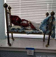 Antique composite doll lying in brass doll bed with bedding in front of window blinds.