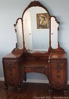 Front view of vintage wooden vanity with triple mirror, detailed carving, and turned legs with casters installed except one missing on back leg.