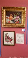 Three framed needleworks hanging on a red wall. Top piece shows jug and fruit still life, bottom left a basket of pink flowers, bottom right a Welcome sign with fruit.