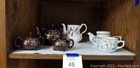 Brown tea pot, creamer, sugar bowl, floral tea pot and floral cup set displayed on wooden shelf with label.