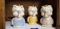 Front view of three ceramic owl pitchers in blue, yellow, and pink with gold accents.