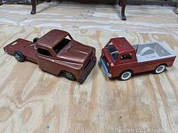 Two vintage toy trucks on a wood floor, one bronze missing the dump bed, one maroon side-gate pickup with white plastic bed.