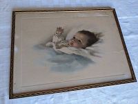 Framed print showing a baby lying with a raised hand, in a gold-tone frame placed on a white textured surface.