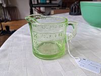 Side and front views of green glass measuring cup showing embossed volume markings in cups and ounces, handle, and pouring spout.