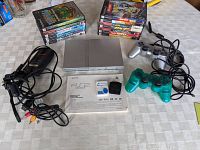 Full lot overview. Silver PS2 slim console, two wired controllers (green and grey), 13 stacked games, cables, memory card, and manual arranged on white checkered tablecloth.