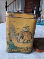 Front side of the tin showing elephant with orange and purple cloth and people around with palace in background