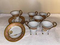 Six footed vintage porcelain cups and six matching saucers with ornate gold detailing and floral patterns on a white background.
