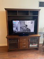 Front view of wooden TV entertainment unit with a TV placed in the opening space. Includes shelves on top, cabinets and drawers on bottom.