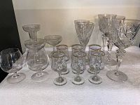 Photo of assortment of 16 various barware glasses showing multiple styles including stemmed sparkling cocktail glasses, floral etched glasses with gold rim, and cut crystal wine glasses.