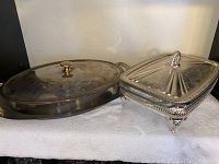 Pair of glass bakeware pieces side by side, showing oval dish with metal handles and lid with brass knob, and rectangular dish with decorative metal lid and ornate metal base with feet.