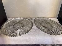 Photo showing a pair of round glass serving trays, one with metal handles and one without, both featuring a radial textured pattern and scalloped edges.