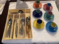 Photo shows 6 colorful espresso cups with matching saucers arranged on a white cloth alongside a plastic cutlery tray holding multiple forks, spoons, and a knife, plus a white ceramic napkin holder behind the tray.