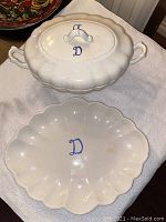 White ceramic soup tureen with lid and blue initial 'D' beside matching serving platter with same initial on scalloped edge platter.