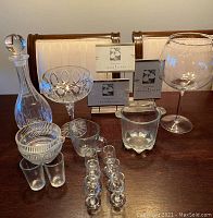 Glass decanter, cut crystal ice bucket, stemware including large goblet and coupe glass, shot glasses on a wooden table with photo displays in the background