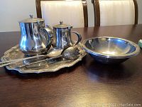 Two silver-tone teapots, one polished round bowl, one decorative bowl, ornate serving tray, and two serving utensils with one being a ladle and the other decorative spoon