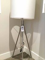 Full view of floor lamp showing tripod base and white fabric drum shade, lamp is standing on carpeted floor against white wall corner.