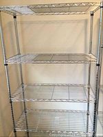 Front view of chrome metal shelving unit showing four of six wire shelves and vertical frame posts, against a beige wall.