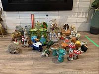 Wide view showing many figurines including frogs with crowns, mushrooms, hedgehog, turtle, bird figurines, and some music boxes on floor against wall.
