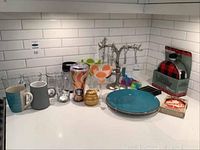 Barware and drinkware items displayed on counter including mugs, glasses, cocktail glasses, plates, canteens boxed set, and small bar accessories.