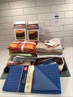 Stack of assorted kitchen linens including folded tea towels, placemats, and two boxed sets of dishcloths with visible colors and textures.