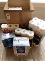 Multiple packs of toilet paper rolls and facial tissues in different brand packaging arranged together around an open cardboard box.