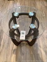 Round side table with clear round tempered glass top held by four metal discs over a dark wood base with three curved intersecting legs.